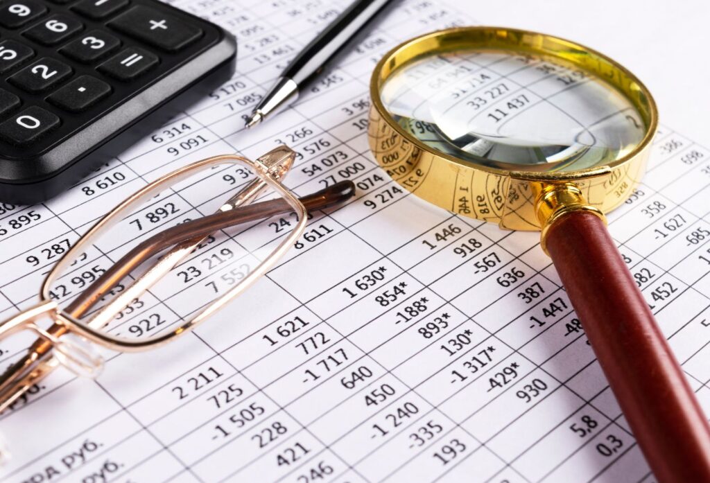 Close-up of financial spreadsheets with numbers, a magnifying glass, glasses, and a pen for analysis/ auditing.”